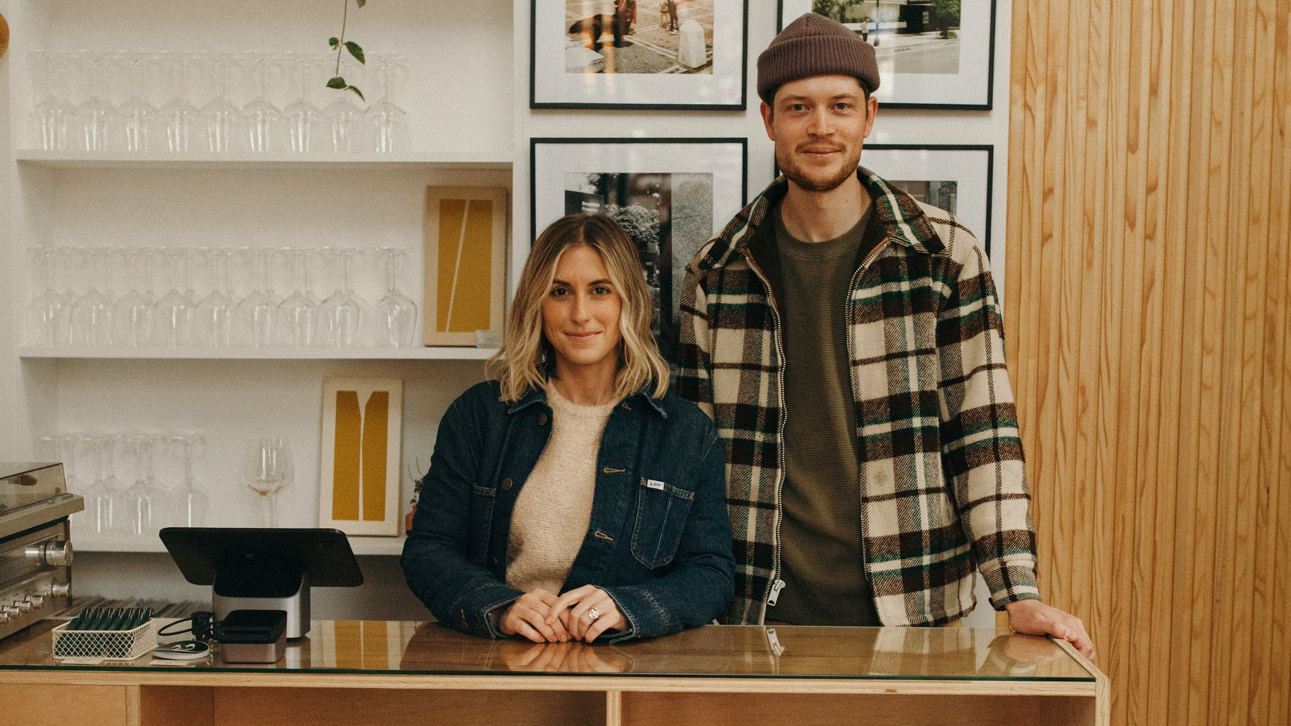 Two people standing behind a bar with shelves stocked with bottles and glasses.